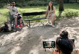 Dreh mit der Hannelore Kohl Stiftung aus Bonn Nele sitzt auf einer Bank und lacht, ich sitze im Rollstuhl und zeige auf Nemo, der neben mir liegt. Wir drei werden von einer Kamera gefilmt.